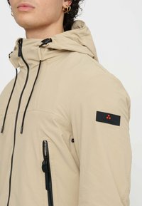 Giacca beige con cappuccio, zip nere e cordini. Presenta una tasca con zip e un'etichetta di marca sulla manica. Tessuto liscio.