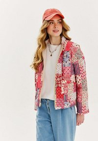 Prešívaná, viacfarebná bunda s kvetinovými a prúžkovanými vzormi, so zipsom a vreckami; nosená cez bielu košeľu a džínsy.