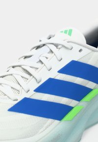 Bijela tenisica s plavim prugama, zelenim detaljima, bijelim vezicama i potplatom sa jastukom, prikazana pod kutom koji ističe bočne i gornje detalje.