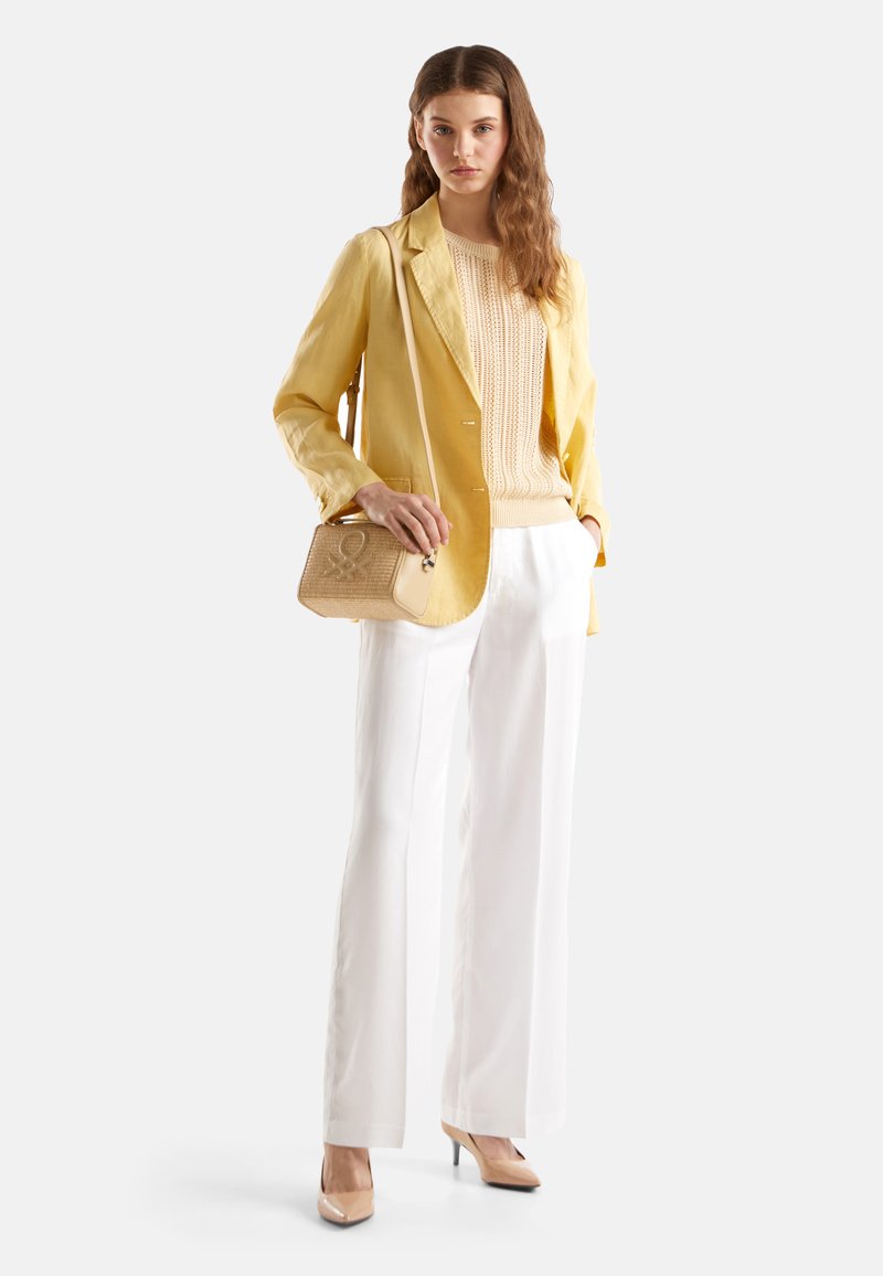 Żółta lniana marynarka na beżowym swetrze z dzianiny, w połączeniu z białymi spodniami o szerokich nogawkach. Trzyma brązową torebkę o sztywnej konstrukcji z detalem logo.
