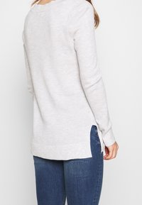 Pullover in maglia grigia chiara con vestibilità rilassata, spacchi laterali e polsini a coste, abbinato a jeans blu scuro.
