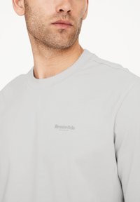 Lysegrå langærmet skjorte med rund halsudskæring og subtilt tekstureret logo på brystet. Blødt stof med en glat overflade.