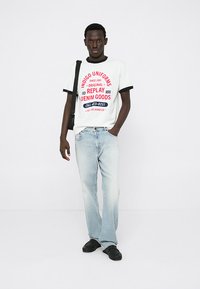 Λευκό μπλουζάκι με γραφικά κείμενα σε κόκκινο και μπλε χρώμα, συνδυασμένο με ανοιχτό μπλε τζιν. Το μοντέλο στέκεται με τα χέρια στη μέση, φορώντας μαύρες παπούτσια και τσάντα ώμου.