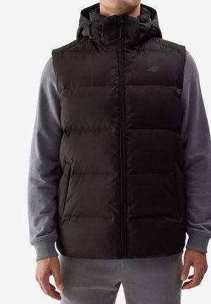 Man met een zwart gewatteerd vest met capuchon over een grijze trui en broek, staand met ontspannen handen langs zijn lichaam.