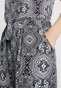 Tissu à motifs noir et blanc présentant des arabesques complexes, des dessins floraux et des formes géométriques. Comprend une ceinture à nouer à la taille et des poches latérales.