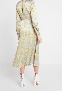 Vaaleansininen midi-mekko, jossa on keltainen leopardikuosi, pitkät hihat, avainaukko selässä ja hulmuhäntä. Yhdistetty valkoisiin nilkkabootseihin.