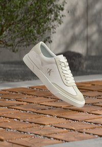 Baskets blanches basses avec le logo "cK" flottant au-dessus d'un sol à motifs de briques et de galets, avec un feuillage vert et un mur gris en arrière-plan.