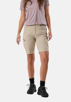 Kvinde iført beige cargoshorts med forlommer, lys lilla t-shirt, sorte sokker og sorte sportssko, stående mod en ensfarvet baggrund.