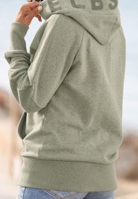 Olijfgroene hoodie met een zachte textuur, voorzien van een verhoogd logo op de kraag en geribbelde manchetten en zoom. Gecombineerd met een blauwe spijkerbroek.