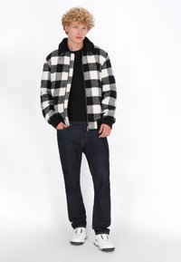 Sort-hvid ternet jakke med sort krave, parret med en sort skjorte og mørke jeans. Modellen bærer hvide sneakers.