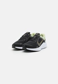 Baskets de sport noires avec tige en mesh, accents vert clair et semelle blanche rembourrée. Présente un logo doré et des lacets noirs.