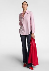 Camicia button-up rosa chiaro a maniche lunghe con colletto a punta e ricamo sottile, abbinata a jeans scuri e giacca di pelle rossa. Scarpe rosse a punta.