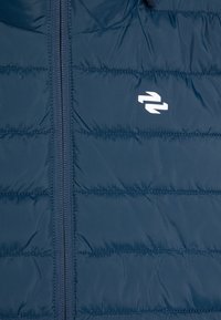 Jachetă de tip puffer navy, cu cusături verticale, fermoar pe toată lungimea și un logo alb pe piept. Material neted, ușor.