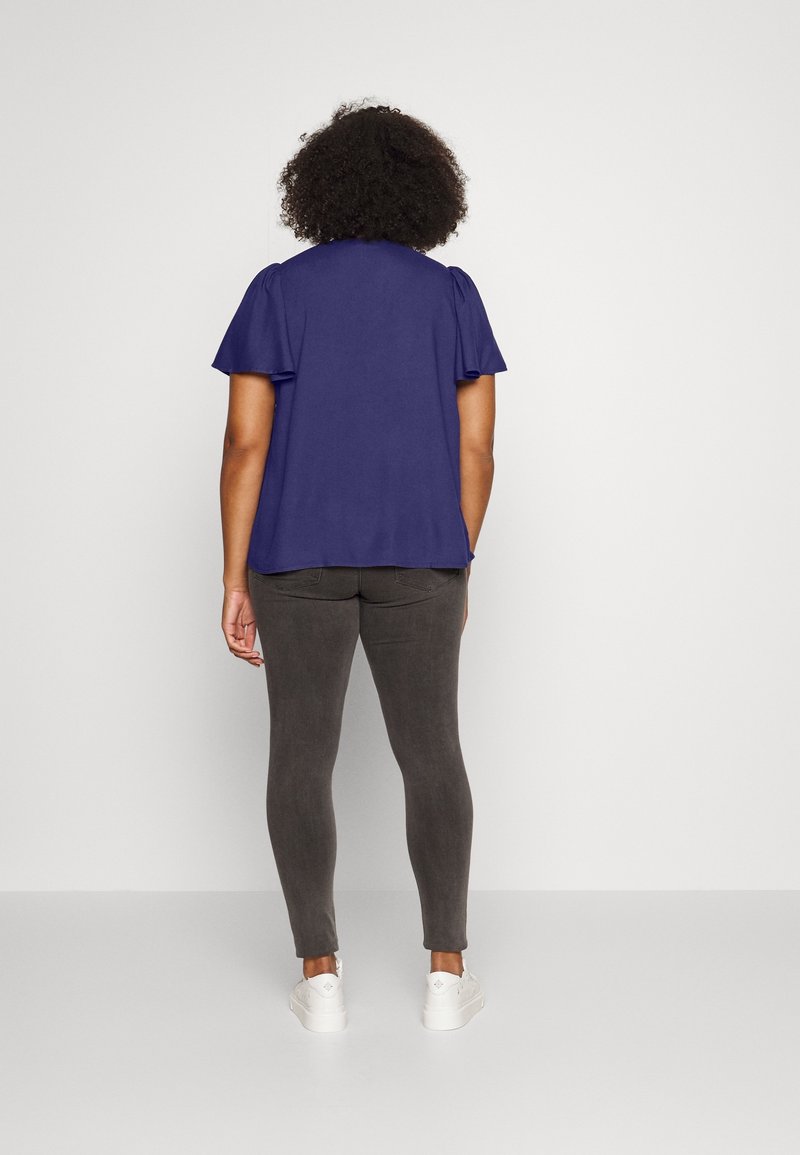 Femme aux cheveux bouclés portant un chemisier violet à manches courtes, un pantalon gris foncé et des baskets blanches, debout de dos à la caméra.