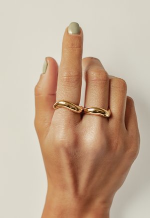 Anillos de oro con un diseño ondulado llevados en dos dedos. La mano tiene un tono de piel clara y presenta un esmalte de uñas verde apagado y corto.