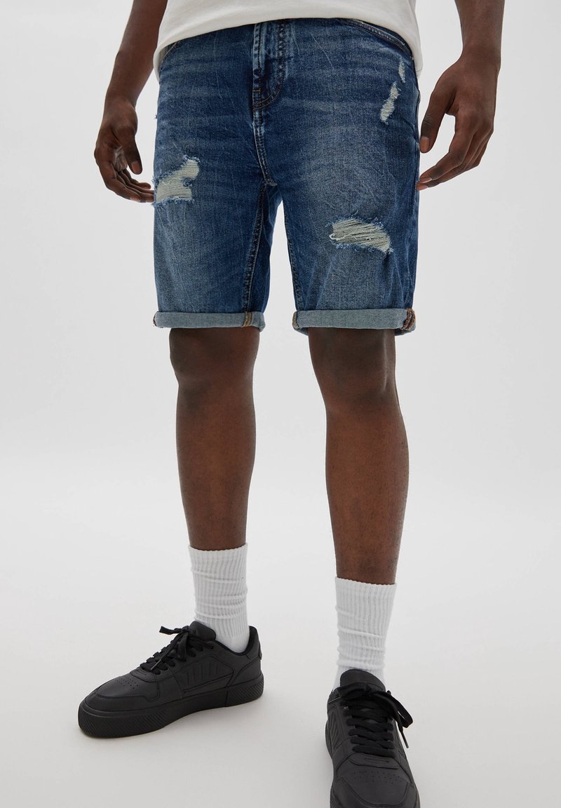 Džínové šortky v tmavě modré barvě s opotřebovanými detaily a ohrnutými nohavicemi. Nosené s černými teniskami a bílými žebrovanými ponožkami.