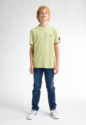 T-shirt vert clair à manches courtes avec col rond et petit logo sur la poitrine, associé à un jean bleu en denim et des baskets blanches.