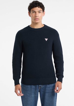 Maglione in maglia a coste di colore blu scuro con maniche lunghe, scollo rotondo e piccolo logo triangolare sul petto a sinistra. Abbinato a jeans blu.