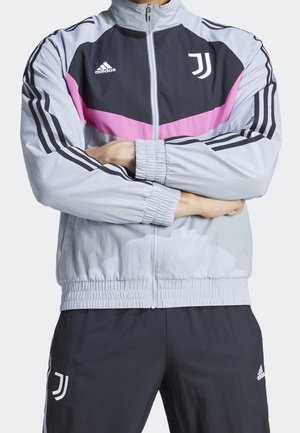 Persona que lleva una chaqueta deportiva Adidas gris claro con detalles negros y rosas y pantalones negros a juego con logotipos de la Juventus, brazos cruzados.