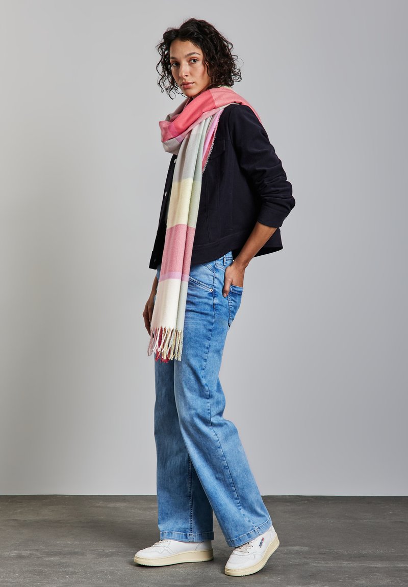 Zachte, multicolore sjaal met roze, gele en witte strepen, gedrapeerd over een zwarte jas. Combinatie met spijkerbroek met wijde pijpen en witte sneakers.