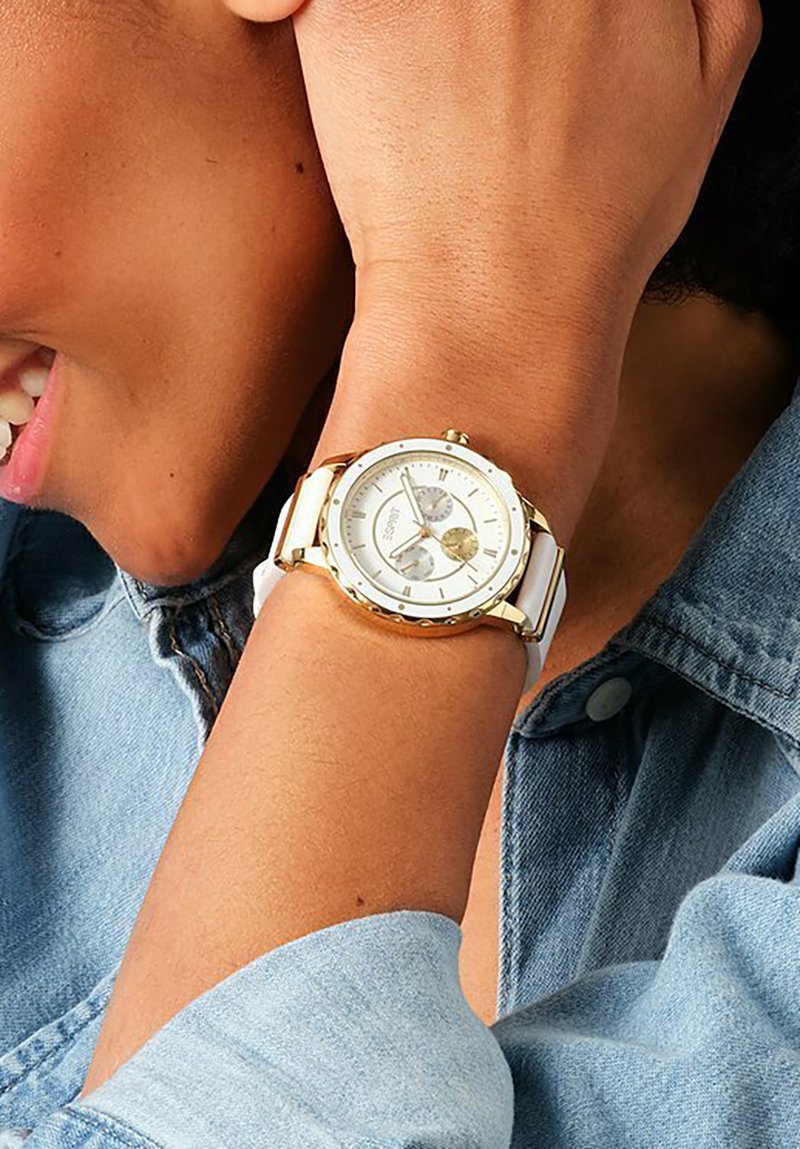Witte horloge met gouden accenten, ronde wijzerplaat, multifunctionele functies en een soepele band. Draagt om een pols, naast denim stof.