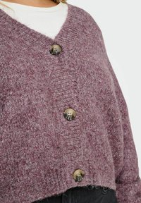 Strickjacke in Helllila mit V-Ausschnitt, ausgestattet mit großen, runden, dunklen Knöpfen und einem lockeren, cropped Design. Texturierte Stoffdetails.