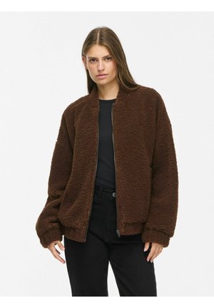 Jeune femme portant une veste zippée duveteuse marron foncé sur un t-shirt noir et un pantalon noir, debout devant un fond blanc uni.