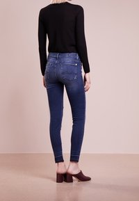 Jeans ajustados en denim azul oscuro con cintura alta, que cuentan con bolsillos traseros y un corte ceñido, combinados con una blusa negra de manga larga y tacones burdeos.