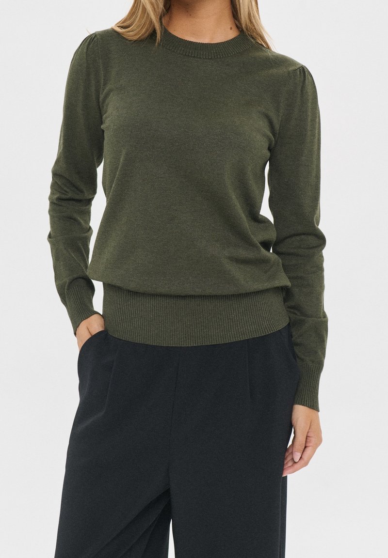 Femme portant un pull à manches longues vert olive avec poignets et ourlet côtelés, associé à un pantalon noir ample, debout avec une main dans la poche.