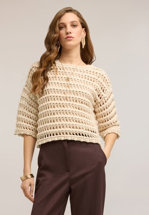 Frau trägt beige offenen Strickpullover, dunkelbraune Hose, goldene Ohrringe, Halskette und Armreifen, steht mit einer Hand in der Tasche.