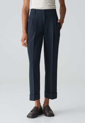 Marineblaue, taillierte Hose mit hoher Taille, zwei vorderen Falten, Seitentaschen und umgeschlagenen Säumen, kombiniert mit dunklen Slippers.
