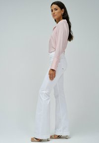 Camisa de manga larga rosa claro con cuello abotonado, combinada con jeans blancos acampanados y sandalias de tacón beige. Texturas suaves y líneas limpias.