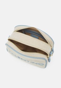 Canvasväska med ljusbeige tyg, blå läderdetaljer, dubbla dragkedjor och en innerficka. Broderad logotyp i blått.