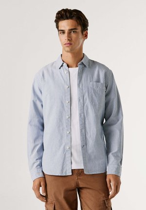 Camisa de botones a rayas en azul claro y blanco, con cuello, bolsillo en el pecho y mangas largas, combinada con pantalones cortos de cargo marrones.