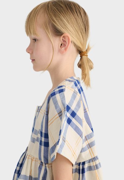 Jeune fille avec des cheveux blonds attachés en une petite queue de cheval, portant une robe à carreaux bleue, blanche et jaune, vue de profil sur un fond uni.