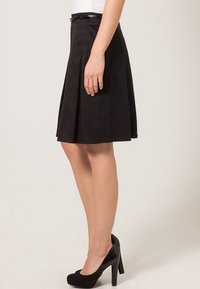 Zwarte plissérok met een aansluitende taille, gemaakt van soepele stof. De zoom valt boven de knie. Gecombineerd met zwarte hoge hakken.
