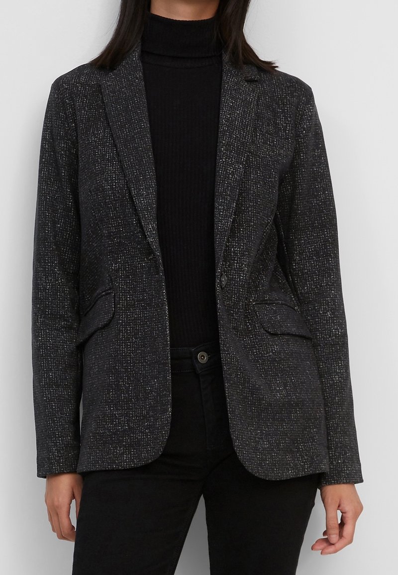 Femme portant un blazer texturé gris foncé par-dessus un col roulé noir et un pantalon noir, se tenant devant un fond clair uni.