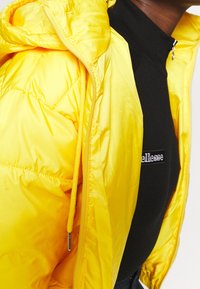 Personne portant une veste matelassée jaune vif par-dessus un haut zippé noir avec un logo blanc partiel sur la poitrine.