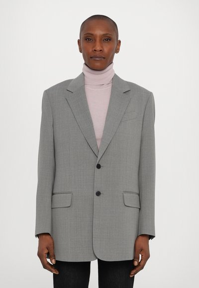 Grau oversized Blazer mit zwei Knöpfen, Reverskragen und aufgesetzten Taschen. Trägt man über einem hellrosa Rollkragenpullover, der Stoff hat eine feine Textur.