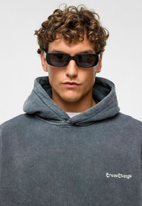 Donkergrijze hoodie met een zachte textuur, voorzien van een voorzak en het "CreateChange"-logo. Het model draagt zwarte rechthoekige zonnebril.
