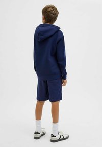 Felpa blu navy con cappuccio, abbinata a pantaloni corti blu navy. Indossata con calze bianche e sneakers con accenti neri. Tessuto morbido, vestibilità casual.