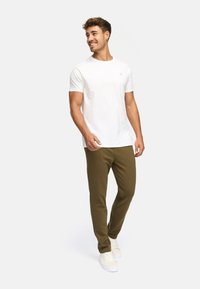 Mann lächelt, geht, trägt ein einfaches weißes T-Shirt, olivgrüne Hosen und weiße Sneakers vor einem einfarbigen Hintergrund.