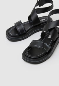 Sandales noires avec de larges brides croisées et une semelle épaisse. Elles présentent une texture lisse, des coutures d'accent bleu et une semelle rectangulaire.
