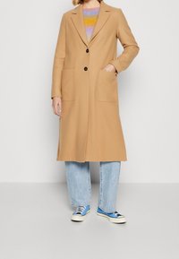 Camelowy długi wełniany płaszcz z kołnierzem, dwoma przednimi kieszeniami i dwoma czarnymi guzikami. W zestawie z jasnoniebieskimi dżinsami i niebieskimi trampkami.