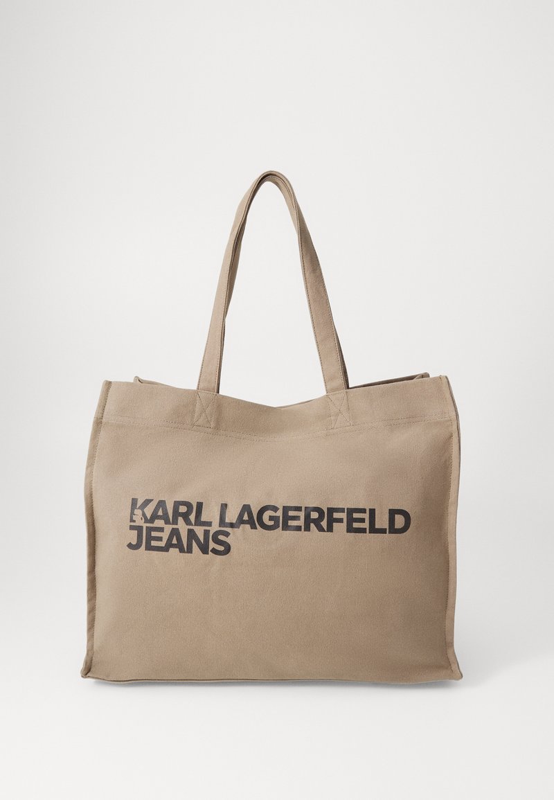 Beige canvas tote bag with dual handles, featuring bold black text "KARL LAGERFELD JEANS" on one side. Simple, unadorned design.