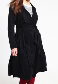 Cappotto avvolgente in suede nero con maniche lunghe, cintura da annodare e due tasche laterali. Il cappotto presenta una texture liscia e un drappeggio frontale.