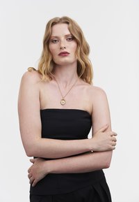 Top nero senza spalline con finitura in rilievo, abbinato a una collana con ciondolo d'oro che presenta un medaglione circolare.