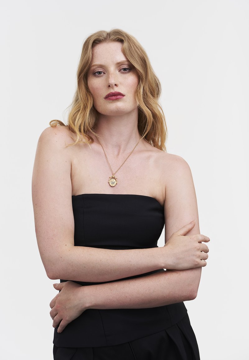 Top nero senza spalline con finitura in rilievo, abbinato a una collana con ciondolo d'oro che presenta un medaglione circolare.