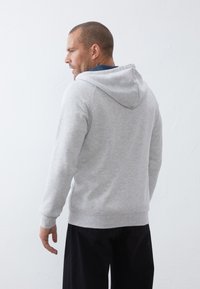 Grijze katoenen hoodie met een trekkoordkap, ribgebreide manchetten en onderrand, in een relaxte fit. Gemaakt in combinatie met donkere broek, getoond vanuit de achterkant.