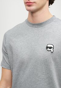 Pilka medvilnės marškinėliai su apvaliu iškirpimu, trumpomis rankovėmis ir juodai baltu grafiniu pleistru kairiajame krūtinės pusėje, vaizduojančiu veidą su akinių nuo saulės.